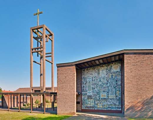 Modernist brick church with an open concrete bell tower and a large ceramic tile mural of saints on its end wall.