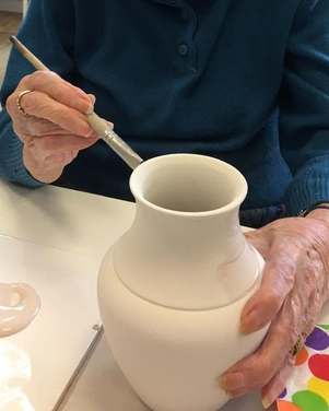 An older woman’s hands, one with a fine brush, the other steadying a plain white bisque vase, beside a palette of pale pink paint