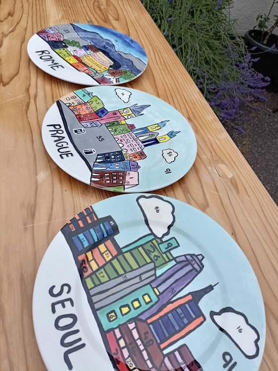 Three hand-painted plates showing colourful city skylines of Rome, Prague and Seoul, laid out on a wooden table outdoors