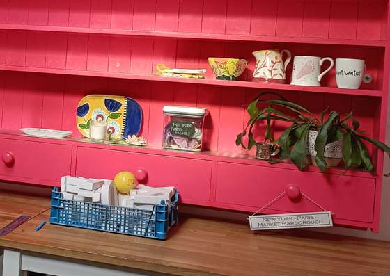 Bright pink dresser with shelves of painted pottery, a pot plant and a hanging sign reading “New York – Paris – Market Harborough”, above a wooden work table