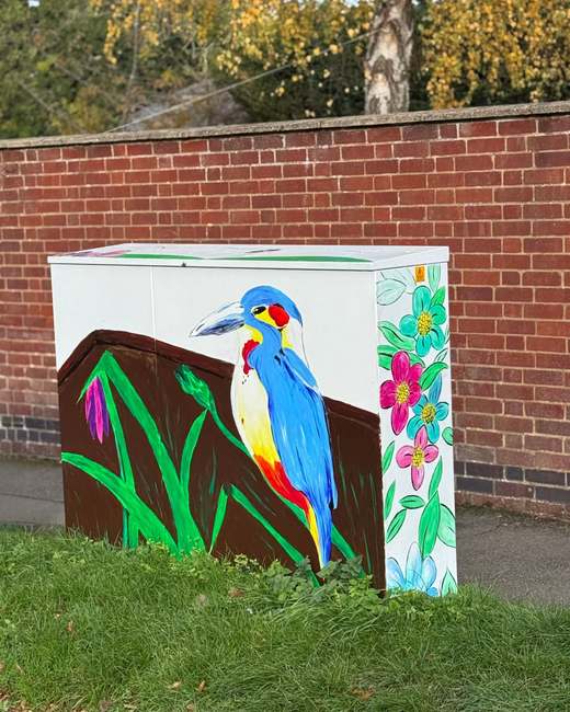 Utility cabinet on Walcot Road painted with a bright blue bird and colourful flowers, in front of a red-brick wall